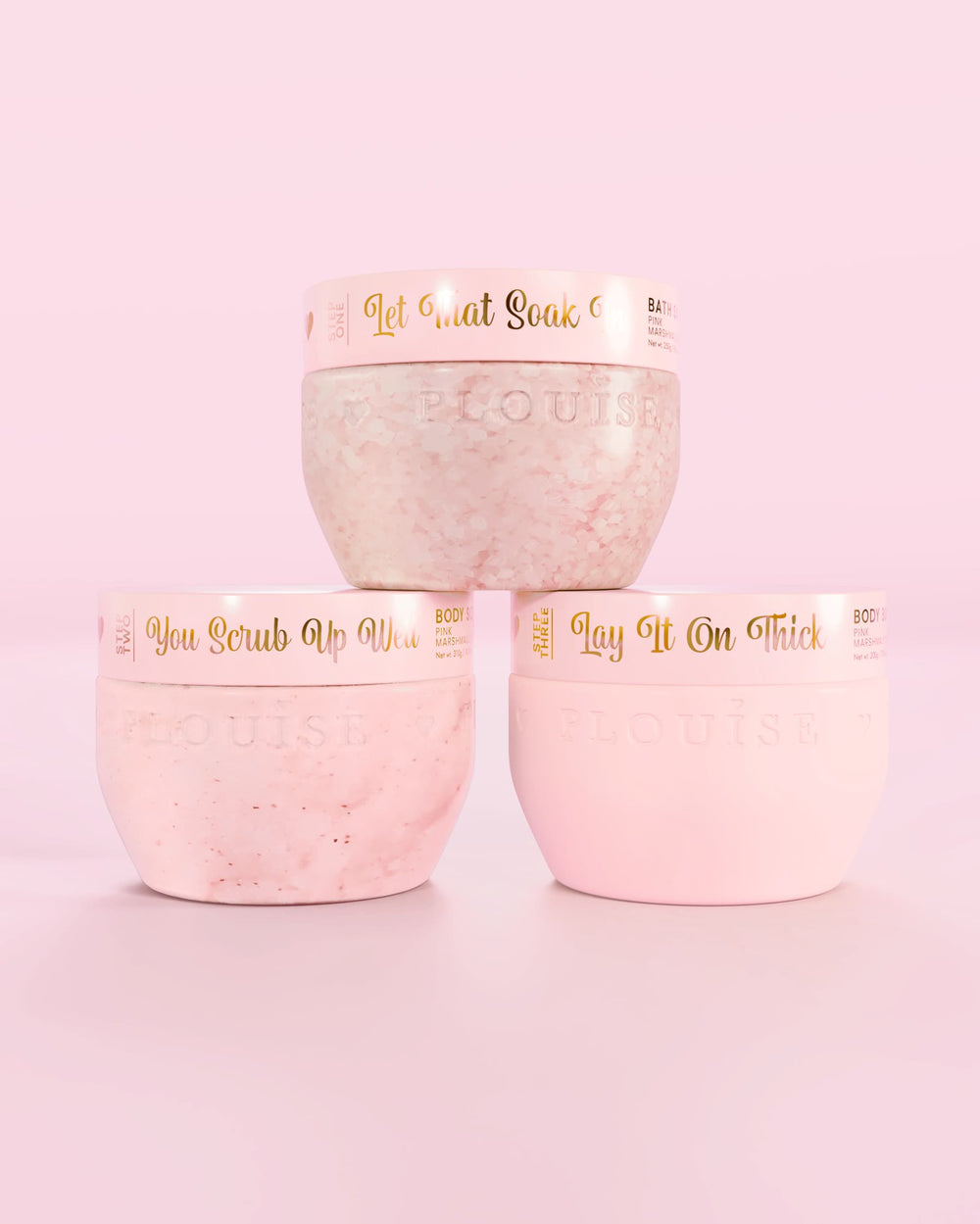 Tři růžové nádobky P.Louise Hop Into Self-Care Pink Marshmallow obsahující koupelové soli, tělový peeling a tělové máslo se zlatým textem na růžovém pozadí