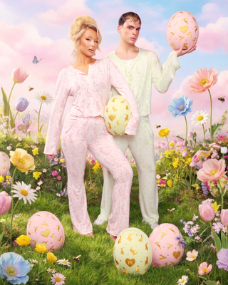 Two people in pastel Easter bunny pyjamas standing in a field of flowers with Easter eggs.