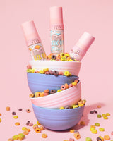 Stack of colourful bowls filled with cereal pouring out and three bottles of the P. Louise Frosted Fix Uo Blurring Setting Sprays in the top bowl on a pink background