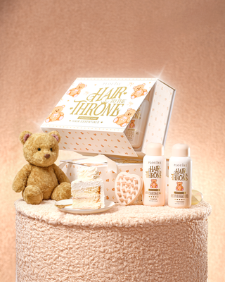 Kuvassa pörröinen pöytä, jossa on P.Louise Hair To The Throne -hiustenhoitosetti, jossa on kookoskakun tuoksuinen shampoo ja hoitoaine sekä päänahan hierontalaite, jonka vieressä on nalle ja kakkuviipale