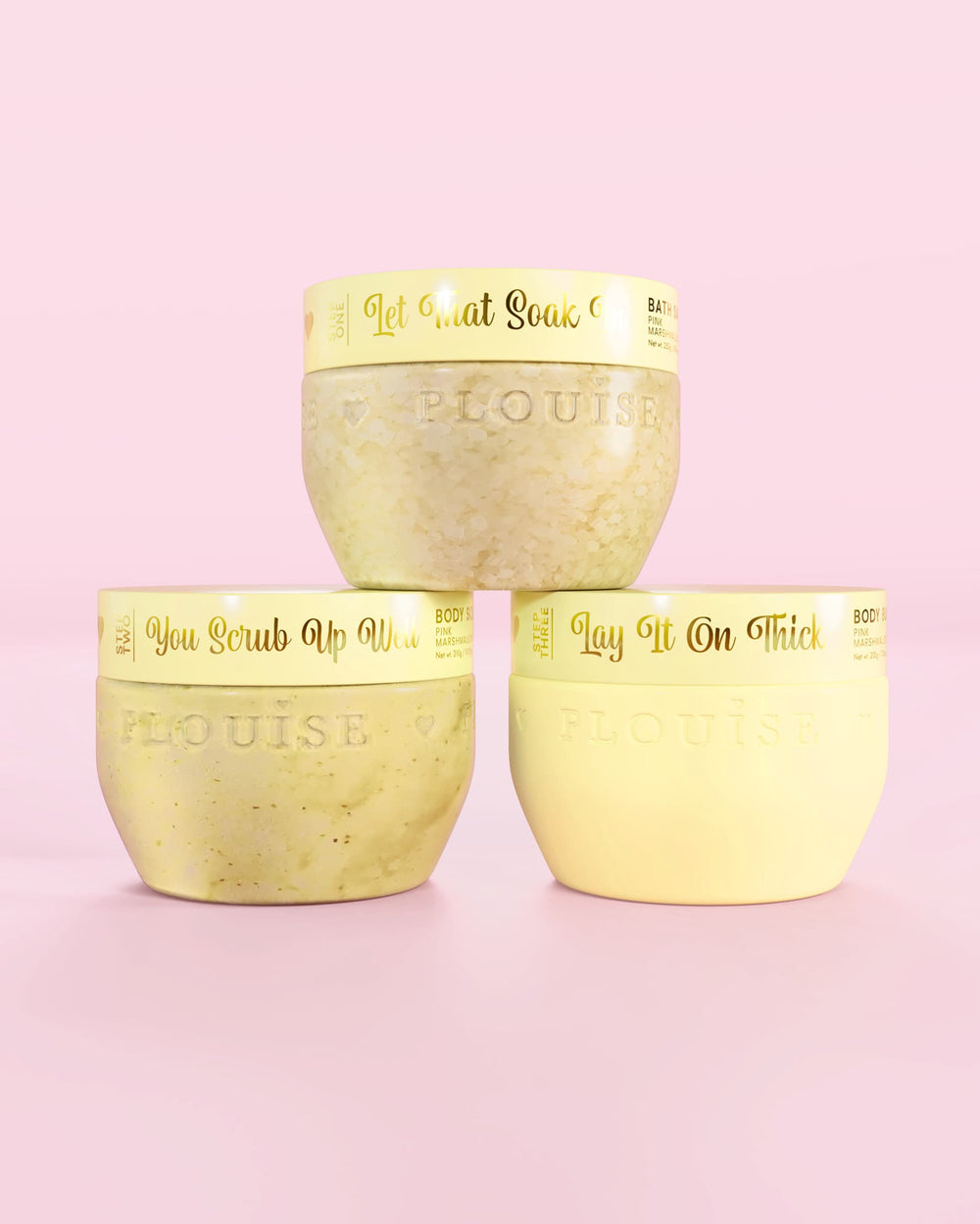 Tři žluté nádoby P.Louise Hop Into Self-Care Starwberry Shortcake obsahující koupelové soli, tělový peeling a tělové máslo se zlatým textem na růžovém pozadí