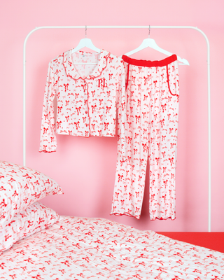 Girls P.Louise julesløyfe pyjamasbuksesett med rød sløyfedesign mot rosa og hvite striper hengende på kleshengere i et rosa soverom med matchende sengetøy