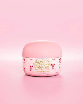 Rosa badekar med SLick It To Me skimmersmeltende kroppsbalsam med dekorative røde sløyfeelementer på en rosa bakgrunn