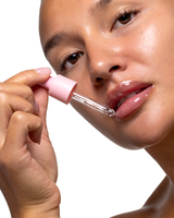 A close-up of a woman applying a clear P.Louise lip serum from a pink dropper tube to her lips against a white background.