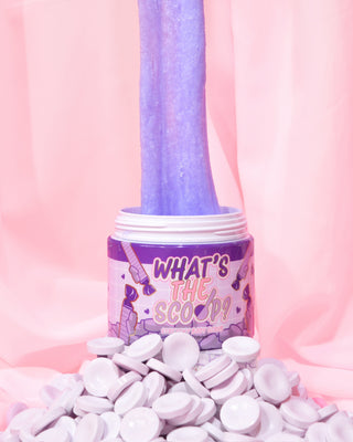 En krukke med 'What's the Scoop?' stretchy bodyscrub af P.Louise vises med en række parma lilla slik i forgrunden, og bodyscrubben strækkes ud af gryden på en lyserød baggrund.