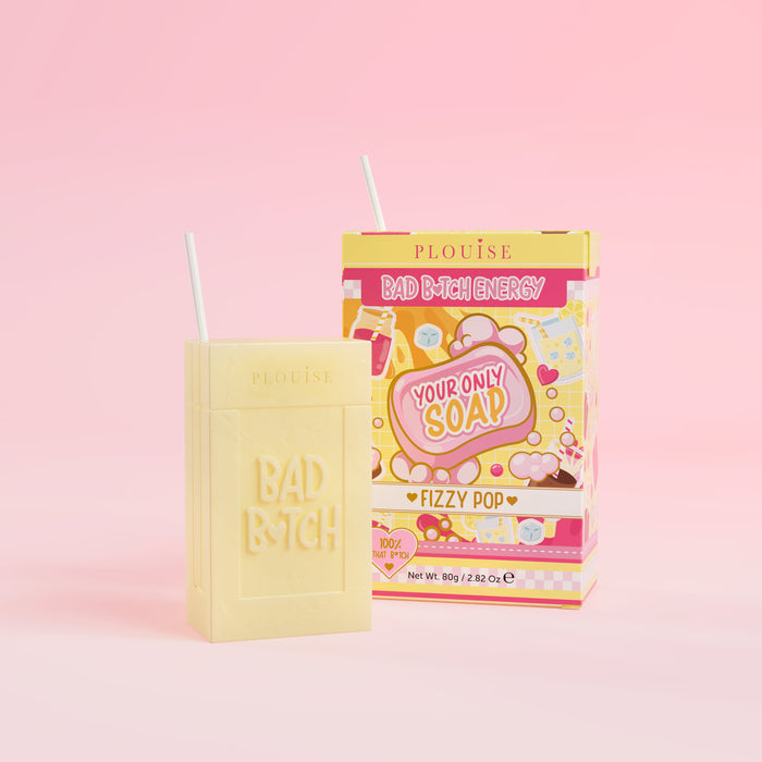 Et gult stykke sæbe med 'Bad Bitch' indgraveret på det, ledsaget af en farverig juicebokspakke med 'Fizzy Pop' mærket på den, præsenteret på en lyserød baggrund.