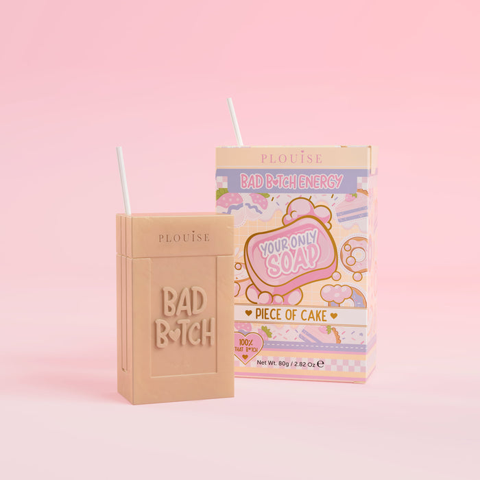 Et beige stykke sæbe med 'Bad Bitch' indgraveret, ledsaget af en farverig juicebokspakke med 'Piece of Cake' mærket på, præsenteret på en lyserød baggrund.