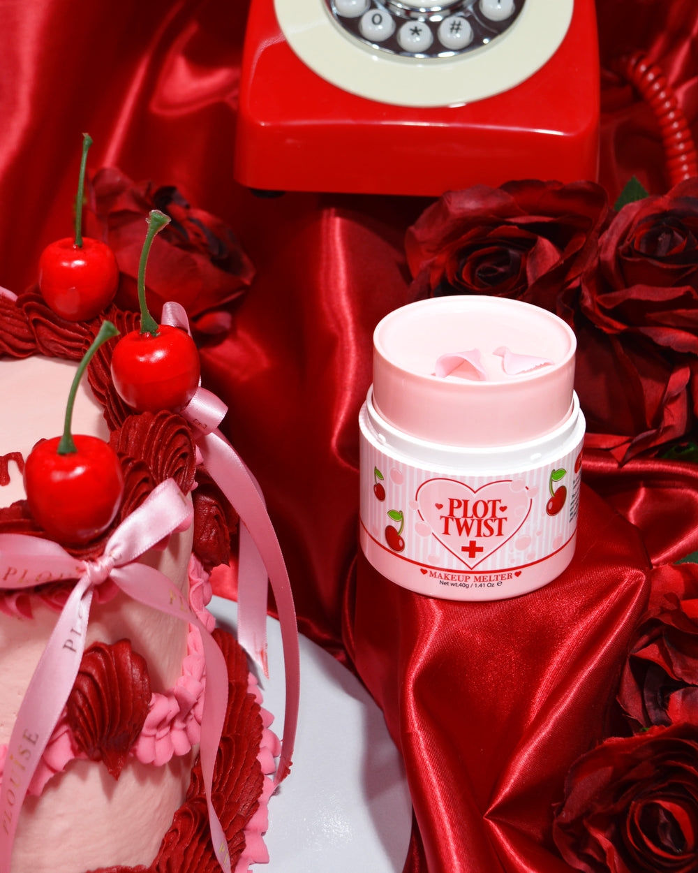 Ένα δοχείο P.Louise Plot Twist Make It Melt Makeup Cleansing Balm που εμφανίζεται σε μια κόκκινη υφασμάτινη επιφάνεια με διακοσμητικά στοιχεία όπως ένα κεράσι και ένα vintage κόκκινο τηλέφωνο στο φόντο.
