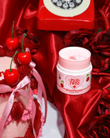 Ένα δοχείο P.Louise Plot Twist Make It Melt Makeup Cleansing Balm που εμφανίζεται σε μια κόκκινη υφασμάτινη επιφάνεια με διακοσμητικά στοιχεία όπως ένα κεράσι και ένα vintage κόκκινο τηλέφωνο στο φόντο.