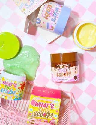 En række farverige strækbare bodyscrub-beholdere med produktnavnet "What's the Scoop?" fremvist markant. Beholderne er arrangeret på en overflade med en lyserød ternet baggrund, og der er legende farverig grafik og tekst knyttet til produktet.